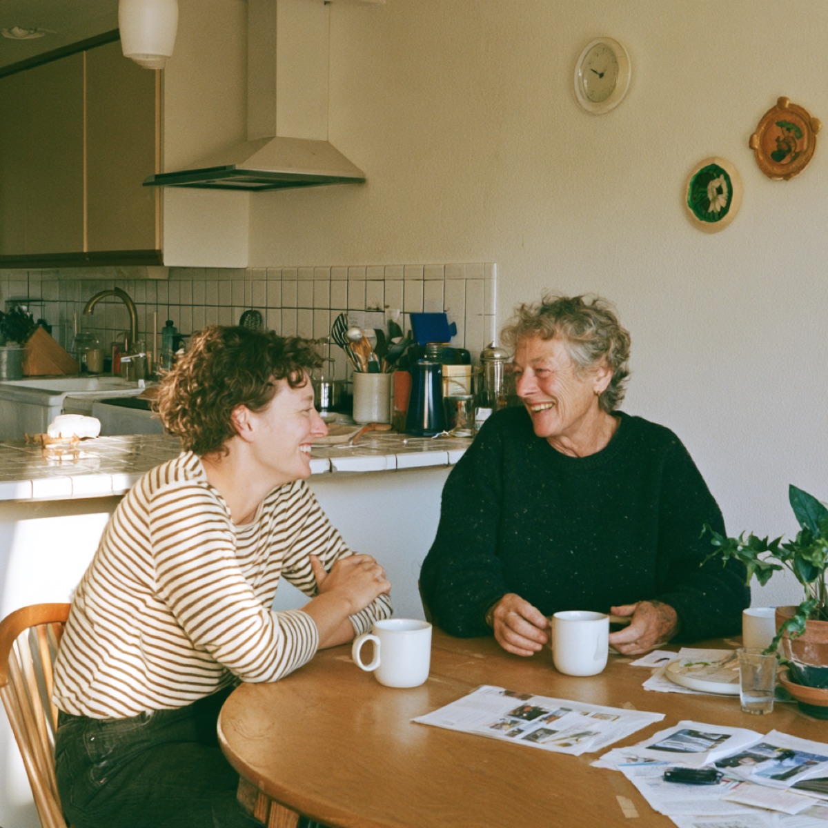 An adult daughter and her elderly mother laughing together over coffee at a sunlit kitchen table